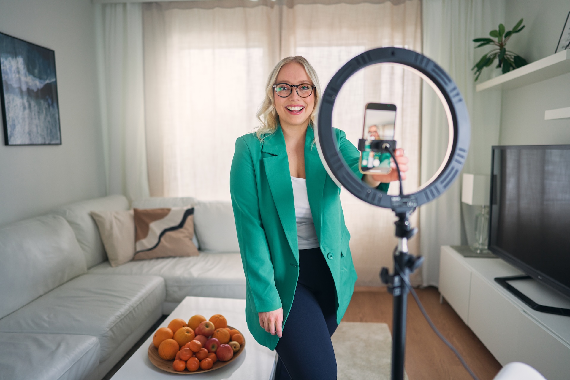 Somestrategi Heini poseeraa hymyillen kameraan p&auml;&auml;ll&auml;&auml;n mintunvihre&auml; bleiseri, edess&auml;&auml;n rengasvalo ja puhelin kuvaamassa h&auml;nt&auml;. Hillitty vaalea tausta kevyine verhoineen.