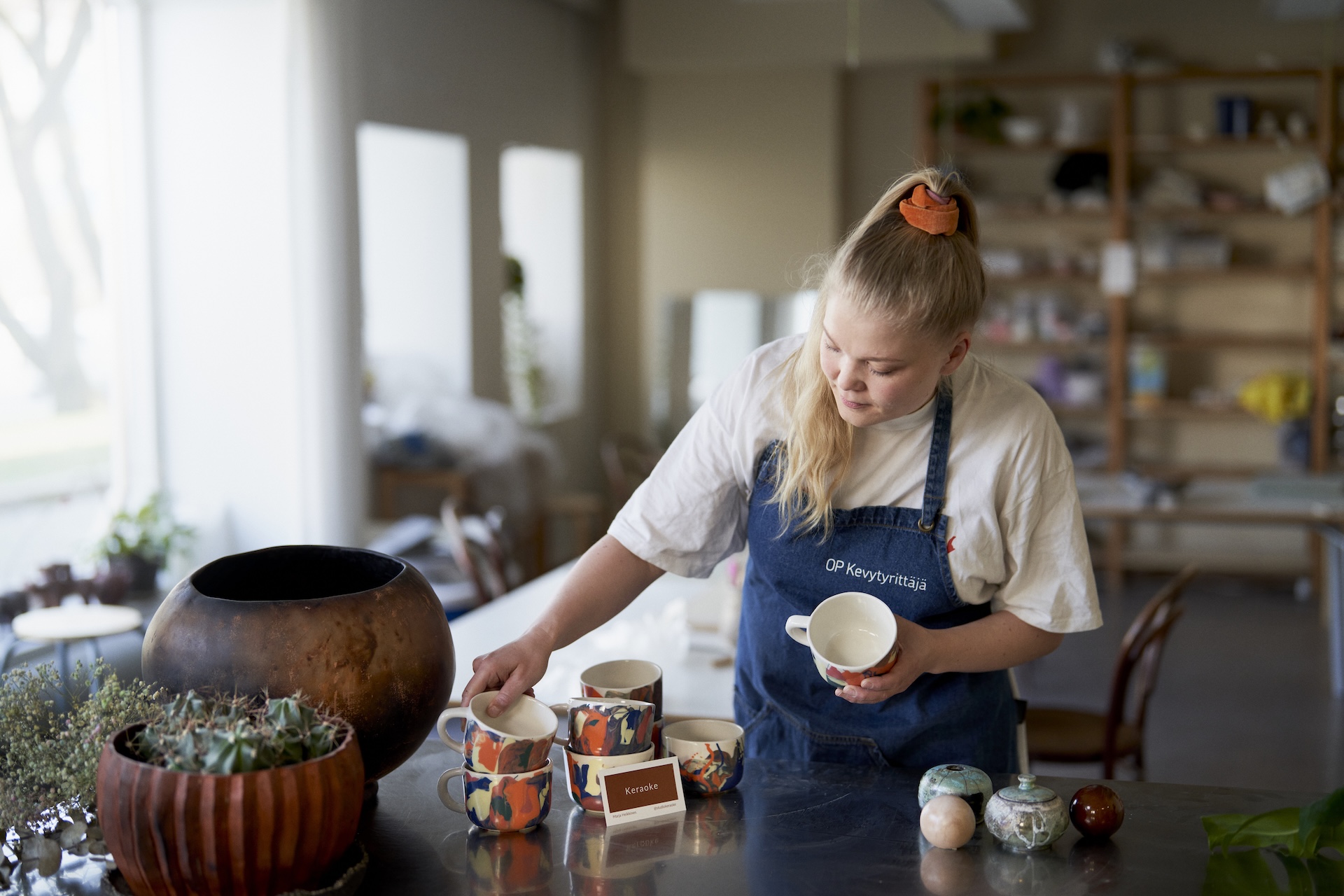 Marja asettelee keramiikkastudiolla kuppeja pöydälle päällään farkkukankainen essu.